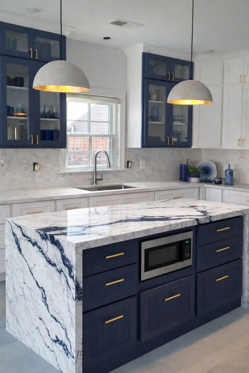 Navy island with a waterfall marble countertop, built-in microwave, glass upper cabinets and dome pendants.