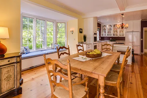 Kitchen and dining room with a central wooden dining table, windows, white cabinetry and hanging lights.
